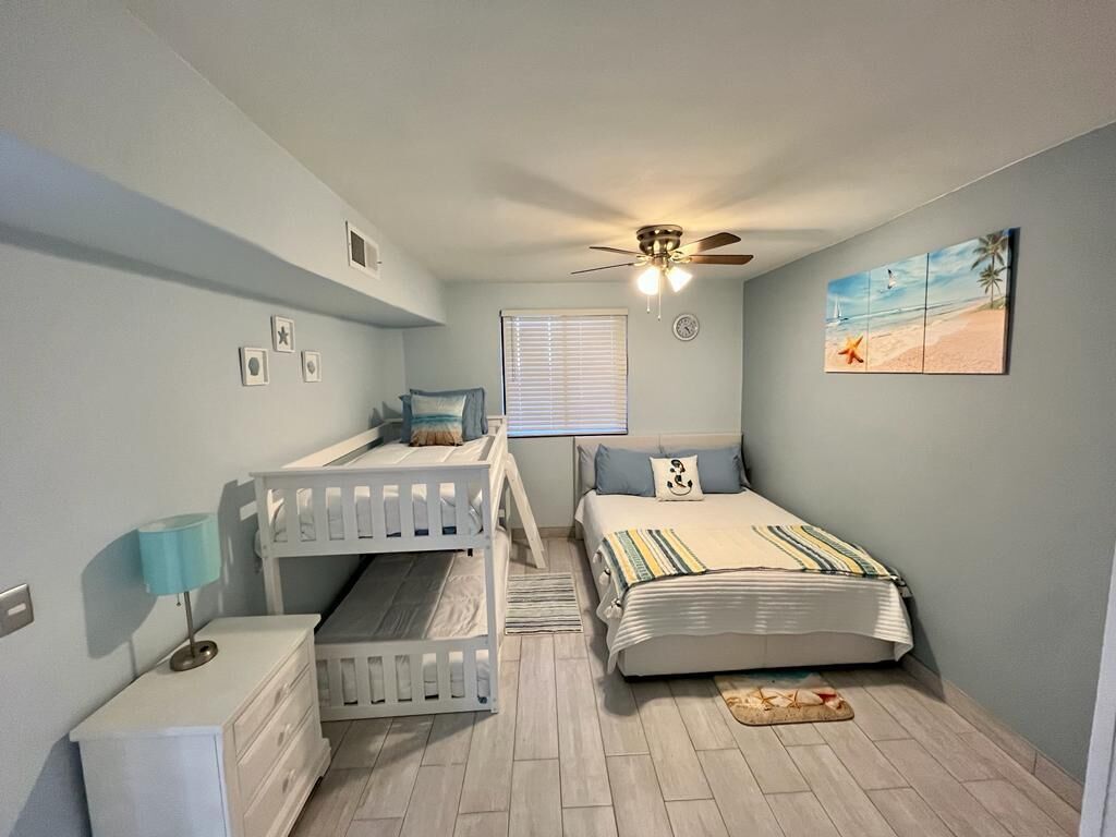 Une chambre avec deux lits superposés, un ventilateur de plafond, un lit avec une couverture à rayures, une table de chevet avec une lampe et un tableau sur le thème de la plage accroché au mur.