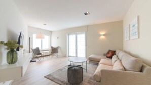 A spacious living area with a sofa, a coffee table, and a dining table. There is a large window providing natural light, and the room has a modern design with minimalistic decor. - Alaska Beach with terrace by Interhome (Bredene)