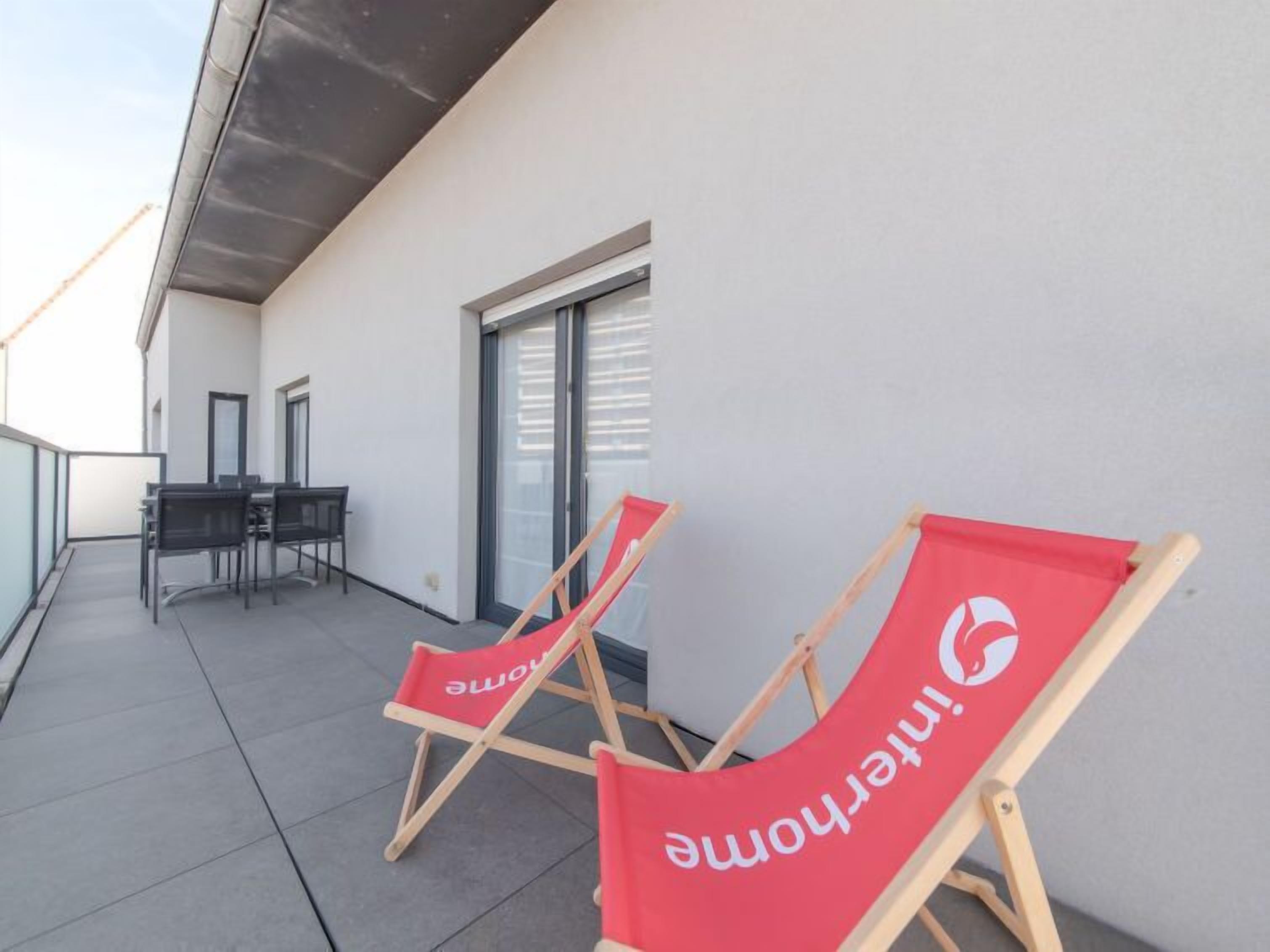 Un balcon avec deux fauteuils rouges et une table avec des chaises, une porte-fenêtre coulissante et un mur blanc.
