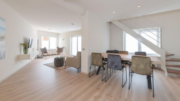 A spacious living area with a modern staircase, a dining table with chairs, and a large window providing natural light. - Alaska Beach with terrace by Interhome (Bredene)