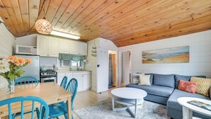 A cozy interior with a wooden ceiling, a dining area with a table and chairs, a kitchen with appliances, and a living area with a sofa and cushions. - 'Tiny House Cottage 5' w/ Crooked Lake Access! (Alanson)