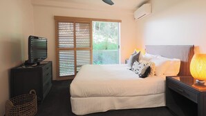 Un dormitorio con cama, televisor sobre una cómoda, ventilador de techo y ventana con persianas.