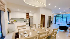 A modern kitchen with white cabinets, a dining table set for four, and a large window providing a view of the outdoors. - Villa 102 Royal Lotus cách bãi biển 5 phút (Hạ Long)