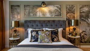 A bedroom with a tufted headboard, a large bed with floral-patterned pillows, two bedside tables with lamps, and framed artwork on the walls. - Views of Palace of the Parliament 3BR Safari Design (Bucharest)