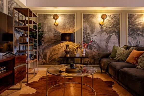 A living room with a sofa, coffee table, and bookshelf. There are wall-mounted lamps and a large painting of palm trees.
