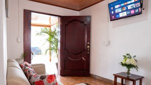 Ein Wohnzimmer mit einer Couch, einer Holztür, einem kleinen Tisch mit einer Vase und einem an der Wand montierten Fernseher, der verschiedene Sender zeigt.