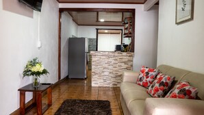 Ein Wohnzimmer mit einem beigefarbenen Sofa, einem dunklen Teppich und einem Holztisch. Im Hintergrund ist eine Küche mit einer Stein-Spritzschutz und einem Kühlschrank zu sehen.