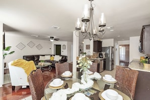 A modern living room with a dining table set for six, a kitchen in the background, and a ceiling fan.
