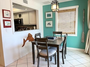 Un coin repas avec une table et des chaises en bois, une cuisine à l’arrière-plan et une fenêtre avec des stores.