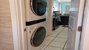 Une buanderie équipée de deux machines à laver superposées, un coin cuisine avec des placards et un plan de travail, et un sol carrelé.