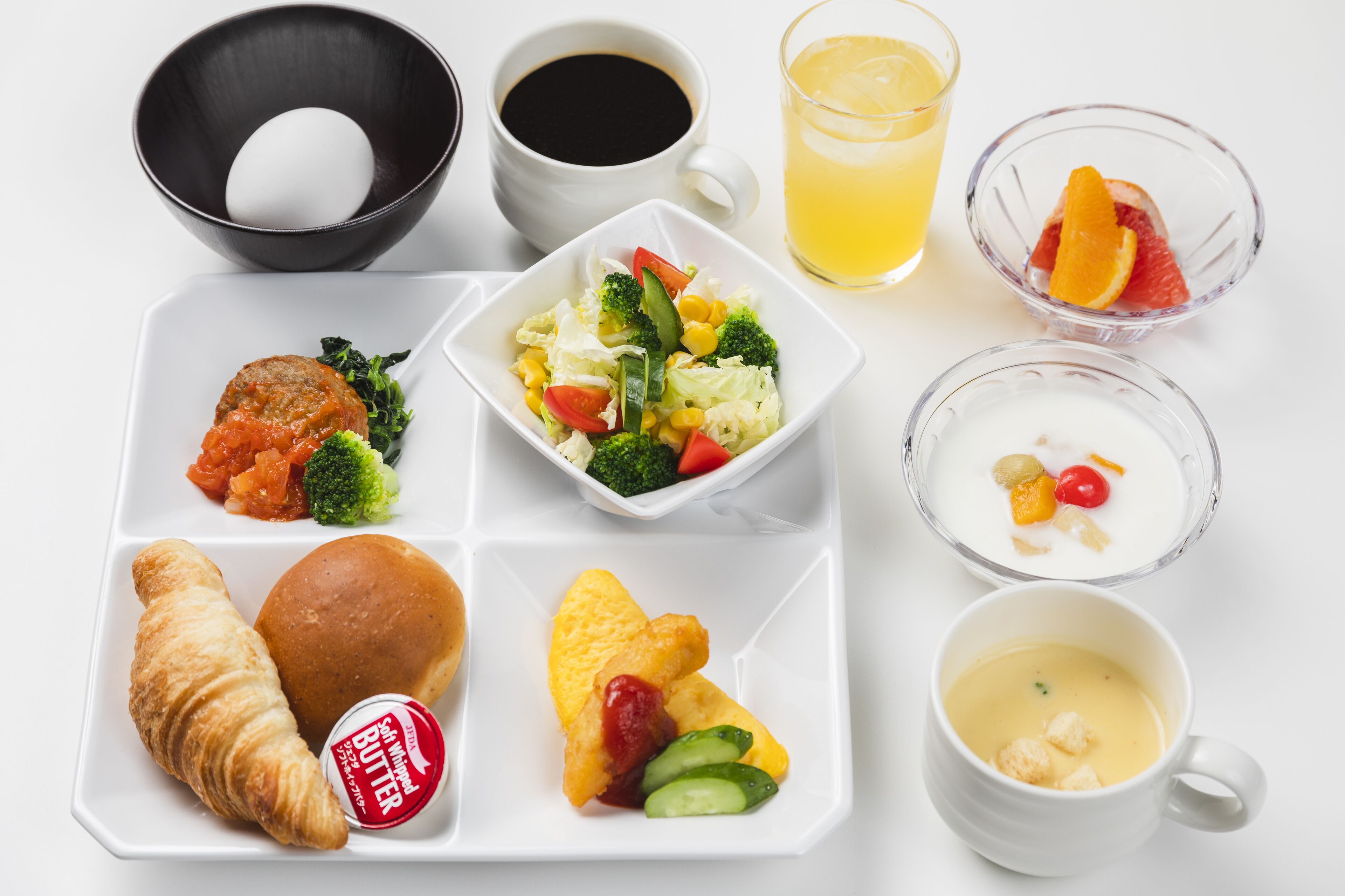 A meal tray with croissants, a small dish of vegetables, a bowl of soup, a glass of juice, and a cup of coffee.