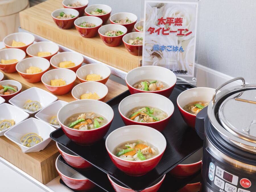A variety of bowls of soup and other dishes on a tray, with a rice cooker and a sign in the background.