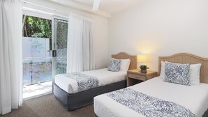 Ein Hotelzimmer mit zwei Betten, einem kleinen Nachttisch mit Lampe und einer Glastür, die zu einem Außenbereich führt.
