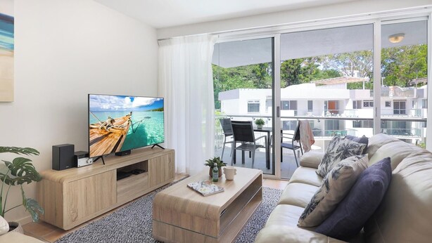 Ein modernes Wohnzimmer mit Flachbildfernseher, Holzmöbeln, einem Glastisch und einem Balkon mit Blick auf Gebäude und Grünflächen.