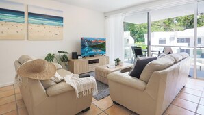 Ein modernes Wohnzimmer mit zwei beigefarbenen Sofas, einem Fernseher, einer Holzkommode und einem großen Fenster mit Blick auf Grünflächen und Gebäude.