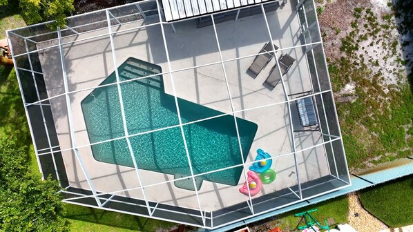 Vista aérea de una piscina con forma geométrica, rodeada por una cerca metálica y equipada con algunas tumbonas y una pequeña mesa.