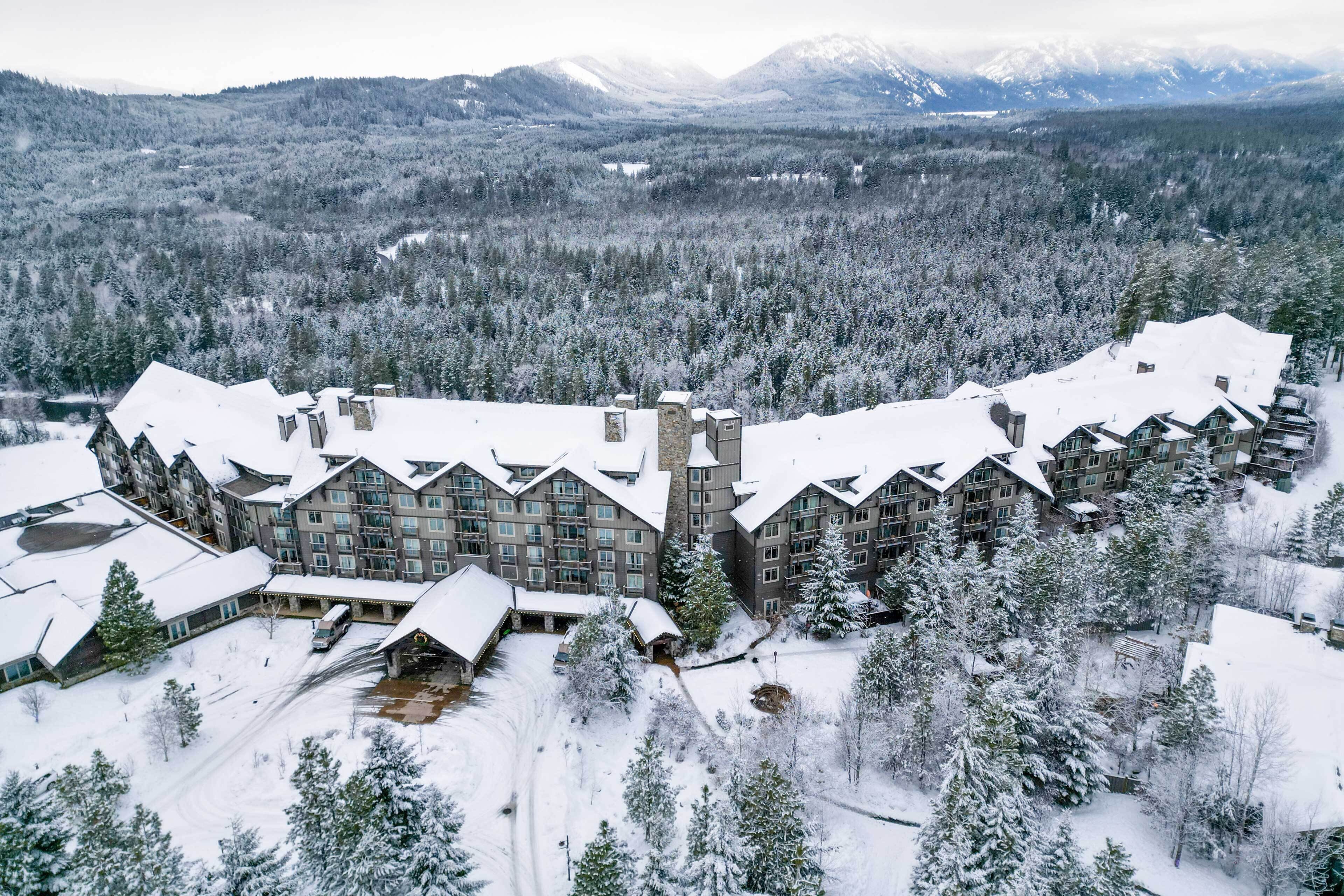 Un grand chalet comprenant plusieurs bâtiments, entouré d’arbres couverts de neige et avec des montagnes enneigées en arrière-plan.