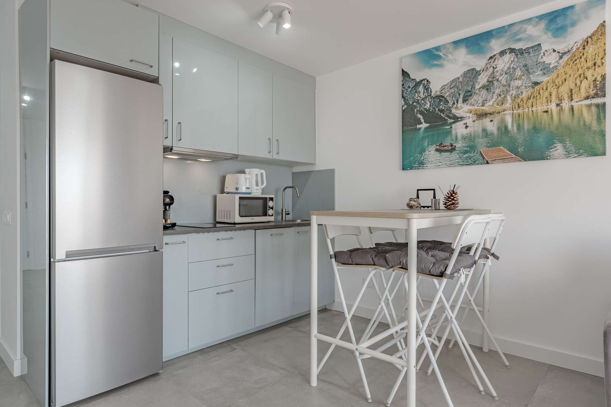 Eine kompakte Küche mit Kühlschrank, Mikrowelle und Kaffeemaschine, ein zusammenklappbarer Esstisch mit Stühlen und ein stimmungsvolles Gemälde einer Berglandschaft an der Wand.
