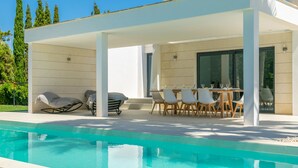 Un área de piscina moderna al aire libre con una zona de estar cubierta, una mesa con sillas y una parrilla.