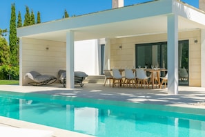 A modern outdoor pool area with a covered seating area, a table with chairs, and a grill.