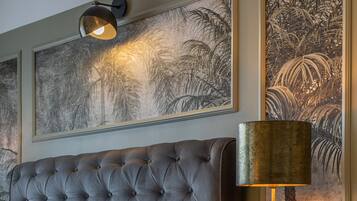 A tufted grey sofa with white and patterned cushions, a round gold table with a lamp, and large botanical wall art.