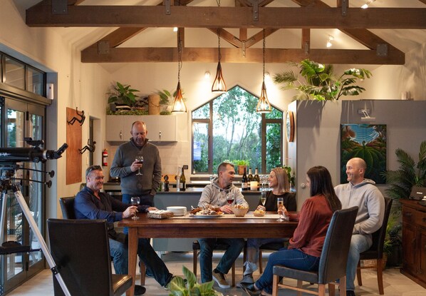 A dining area with a wooden table, chairs, and hanging pendant lights. There are potted plants and a window with a view of trees.