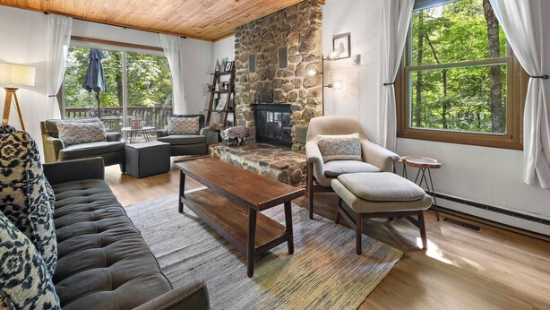 Un salon chaleureux avec une cheminée en pierre, des sièges confortables et de grandes fenêtres donnant sur l’extérieur.