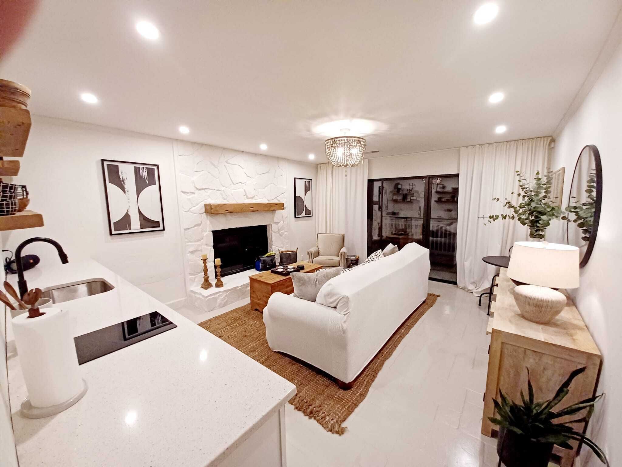 A modern living room with a white sofa, a fireplace, a kitchen area with a sink, and a dining table.