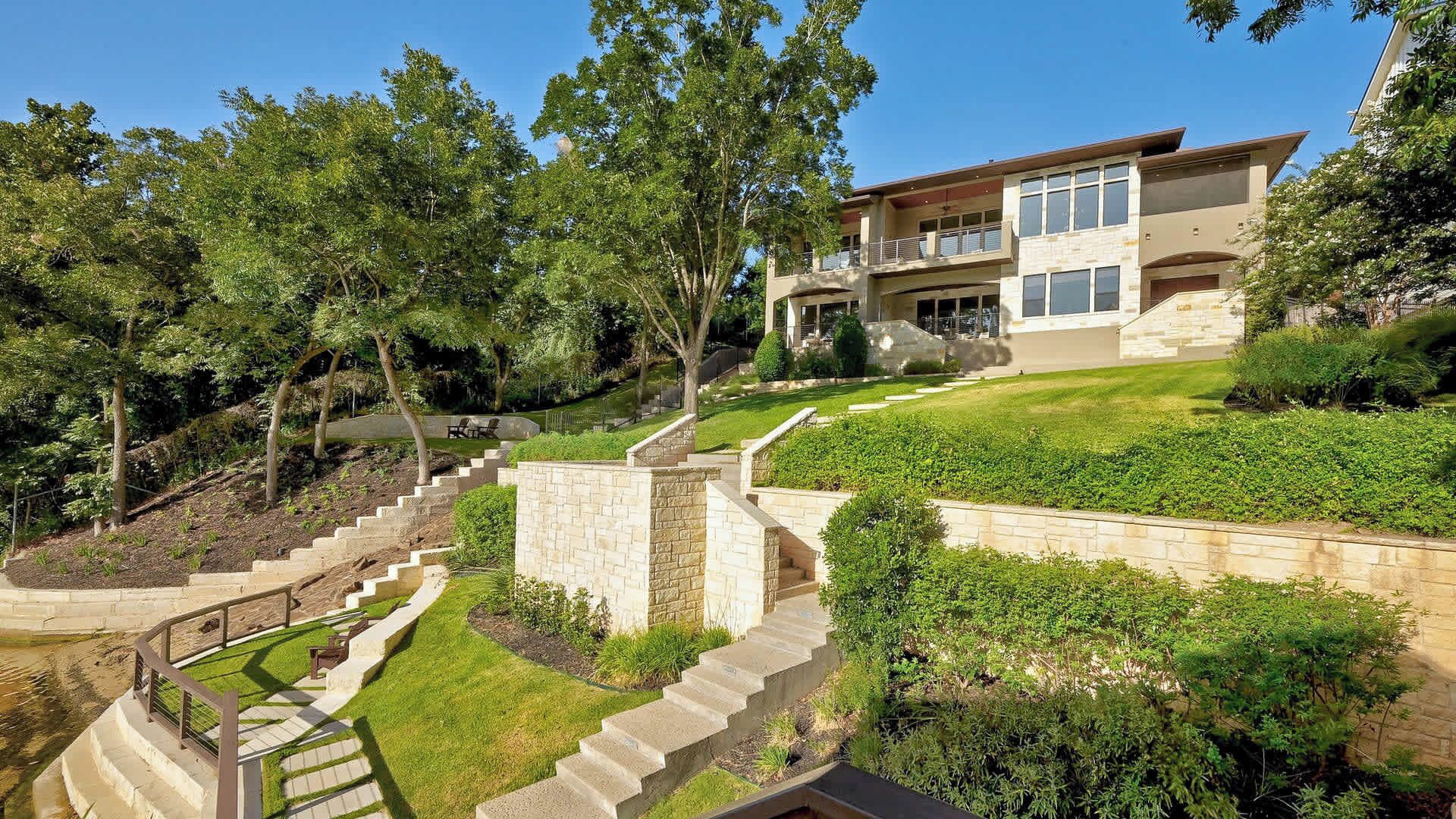 A modern two-story house with a stone staircase leading to the entrance, surrounded by lush greenery and trees.
