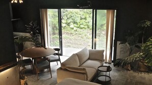 A modern living room with a sofa, a round table, and chairs. There is a large window with a view of greenery outside. - LAVA SAUNA & VILLA (Hachijo)