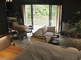 Una sala de estar moderna con un sofá, una mesa redonda y sillas. Se aprecia un ventanal con vistas al exterior, lleno de vegetación.