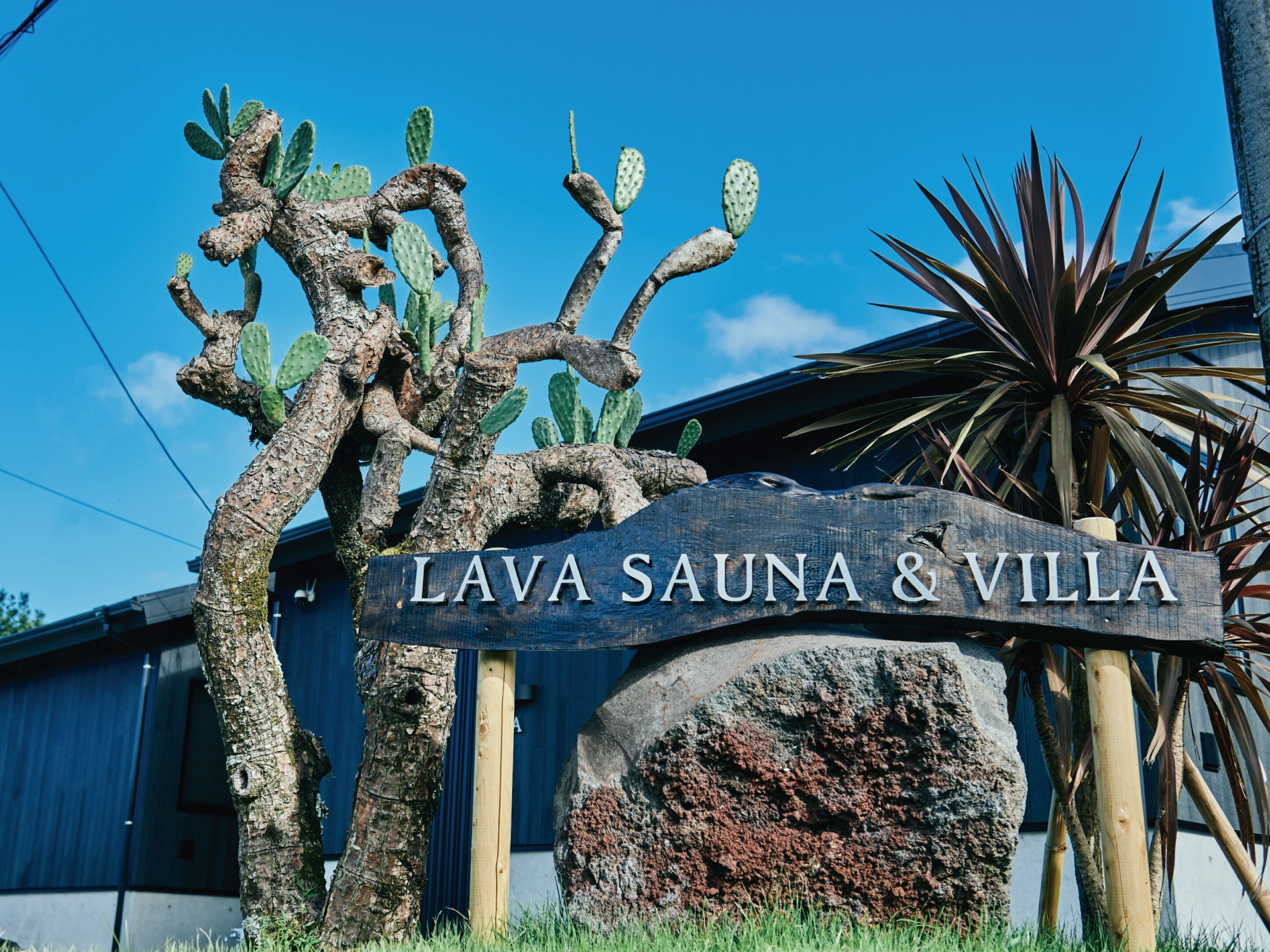 Een bordje voor Lava Sauna & Villa, met een rustiek houten ontwerp, omringd door cactussen en een palm.