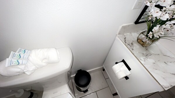 A bathroom with a toilet, a trash bin, a toilet paper holder, and a marble countertop with a vase of flowers.