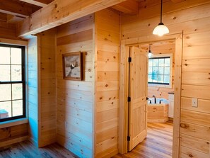 Een houten blokhut van binnen met een gang naar een badkamer, een lijst met een afbeelding aan de muur en een raam met uitzicht.