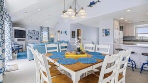 Een ruime eethoek met een houten tafel en witte stoelen, een keuken met witte kastjes, en uitzicht op zee.