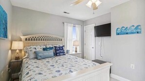 Een slaapkamer met een bed, nachtkastjes, een plafondventilator, een televisie en een raam met gordijnen.