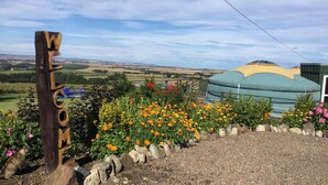 Un letrero de madera con la palabra 'WELCOME' tallada, rodeado de flores de colores y con vistas a una estructura tipo tienda de campaña verde en un paisaje rural.