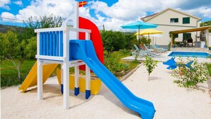 Ein Spielplatz mit Rutsche und einer Flagge, ein Poolbereich mit Liegestühlen und ein Haus im Hintergrund.