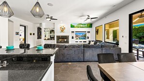 Una cocina moderna con una isla grande, un comedor con mesa y sillas, y una sala de estar con sofá y sillones. Hay un ventilador de techo, luces colgantes y un letrero que dice 'Hollywood'.