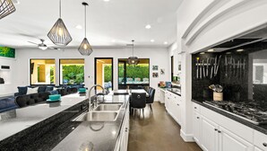 Una cocina moderna con una isla central, encimeras negras, armarios blancos y una estufa negra. Se observan luces colgantes, un ventilador de techo y vistas a la vegetación exterior.