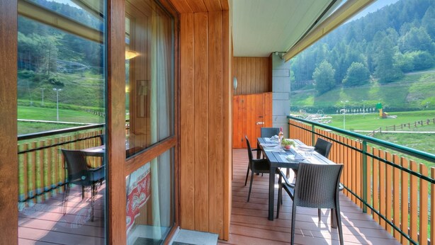 Varanda com mesa e cadeiras, vista para uma área arborizada e guarda-corpo de madeira.