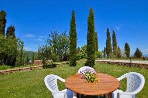 A round wooden table with white plastic chairs, a floral centerpiece, and a clear view of tall trees and a distant landscape. - Villa La Chiesina, Arcidosso, Italy (Arcidosso)
