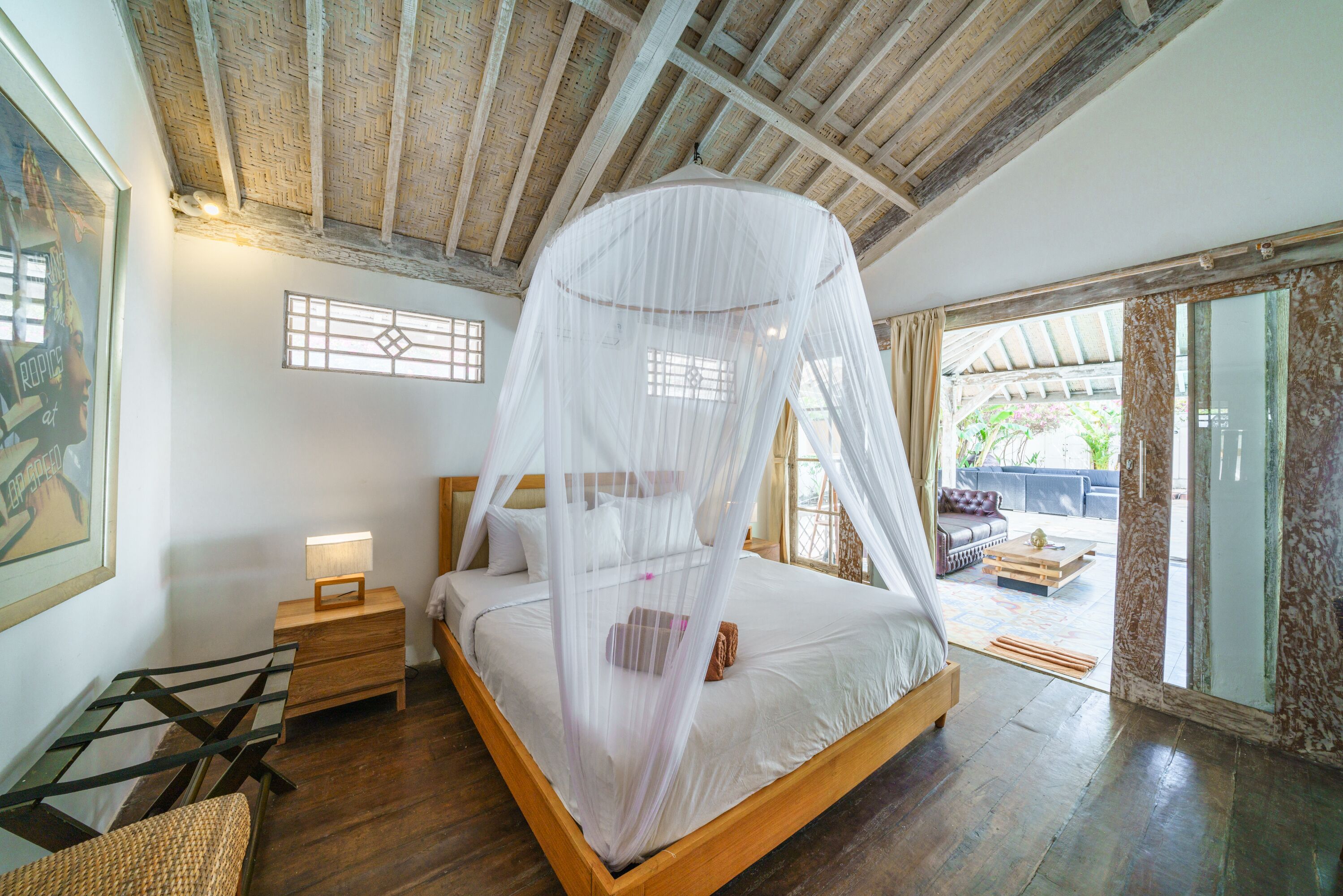 A bedroom with a wooden bed, a nightstand, a bedside table with a lamp, and a large window with a view of the outdoors.