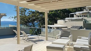 Un espacio al aire libre techado con sillones cómodos y mesas, con vistas panorámicas al mar y a los árboles.