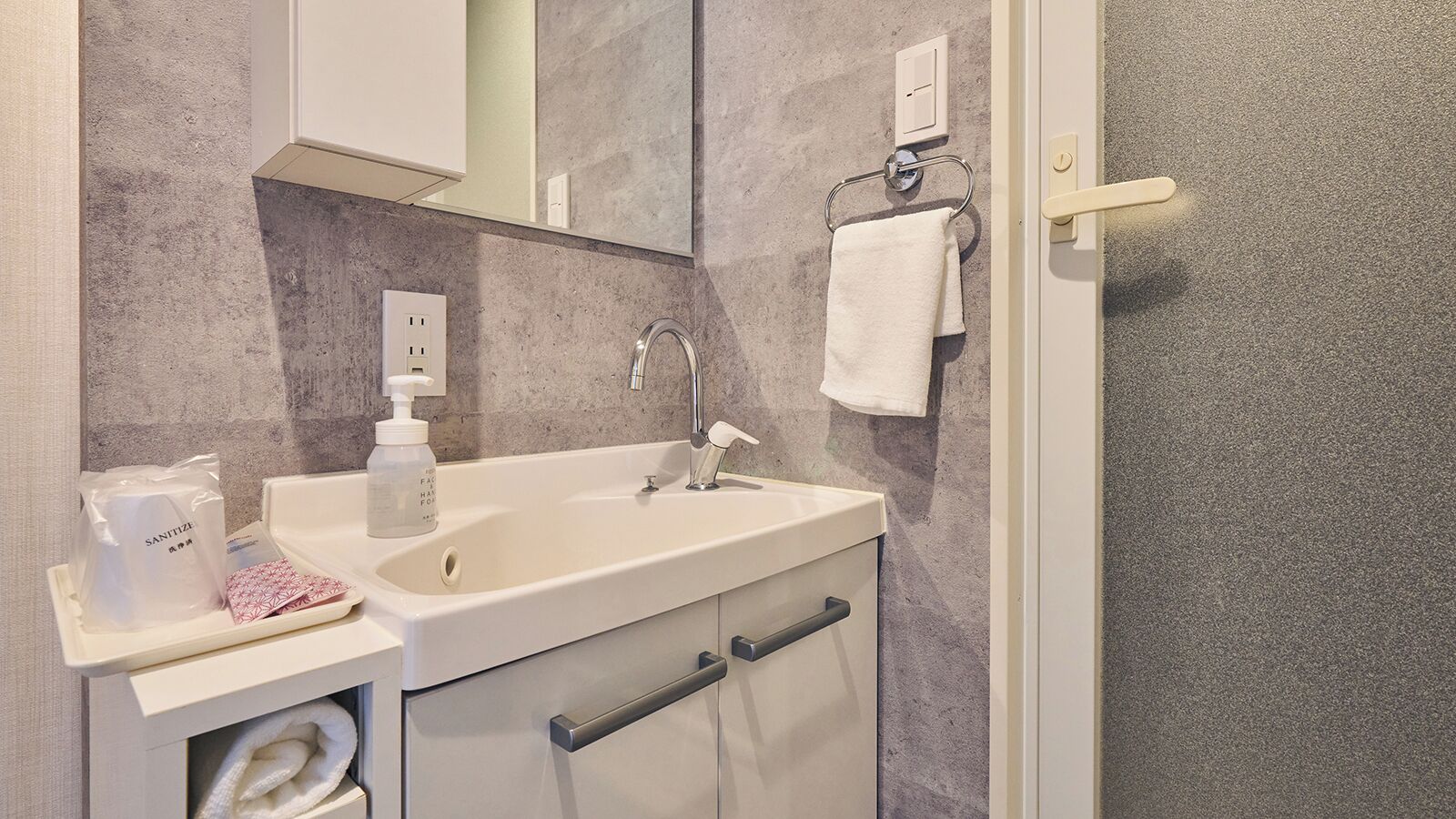 Un baño con un lavamanos blanco, un espejo encima, un perchero con una toalla blanca y un dispensador de gel antibacterial en la encimera.