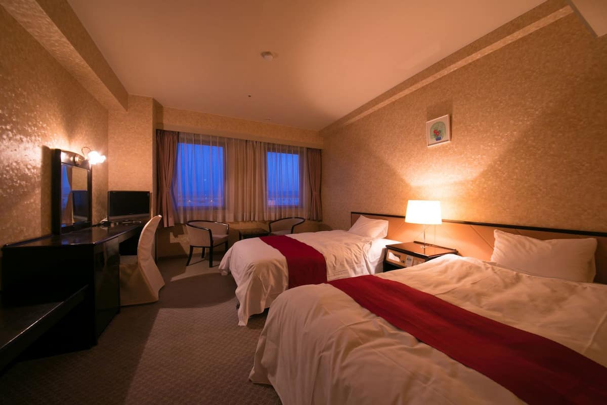 A hotel room with two beds, a desk, a chair, a TV, and a window with curtains.