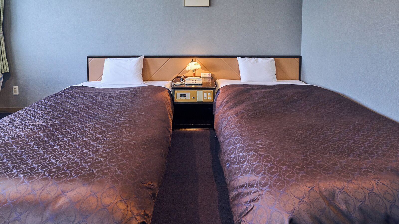 Una camera d'albergo con due letti singoli, ognuno con una coperta a motivi decorativi, un comodino con una lampada e un telefono fissato al muro.