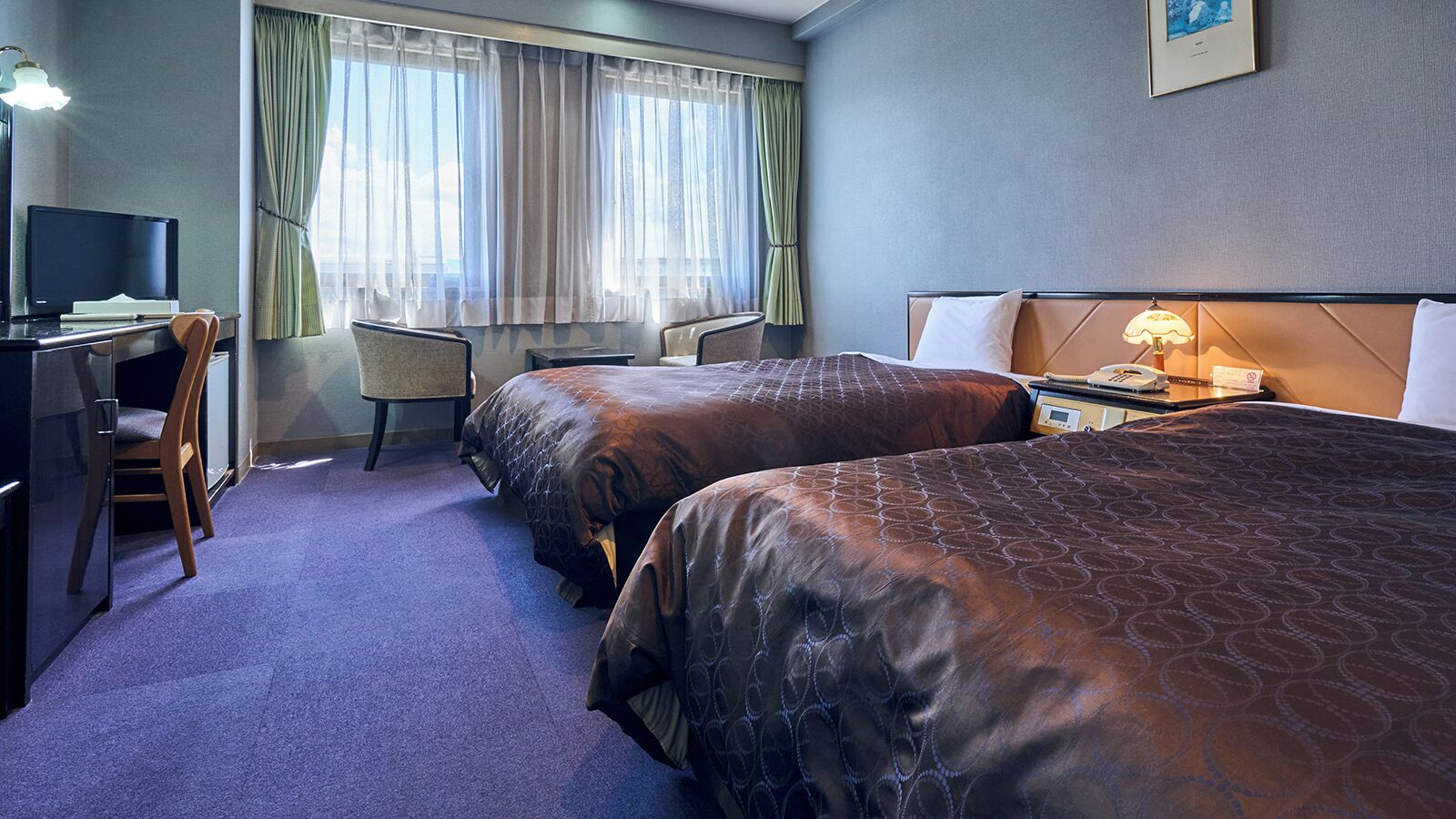 A hotel room with two beds, a desk with a computer, a chair, and a bedside table with a lamp.