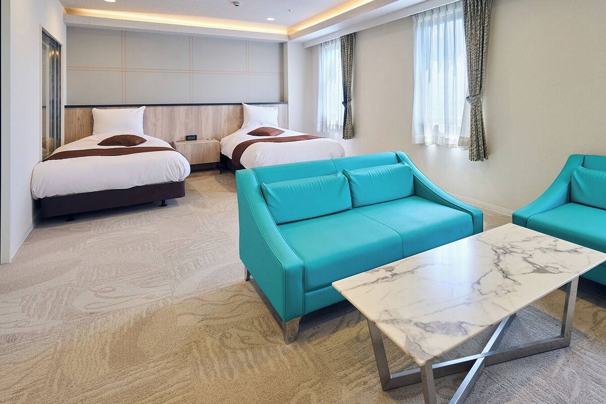 A hotel room with a bed, two teal sofas, a marble coffee table, and a large window with sheer curtains.
