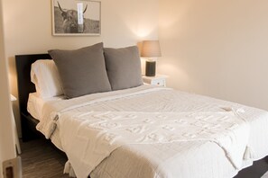 Ein ordentlich bezogenes Bett mit grau-weißer Tagesdecke, zwei Kissen und einer Nachttischlampe auf einem weißen Nachttisch.